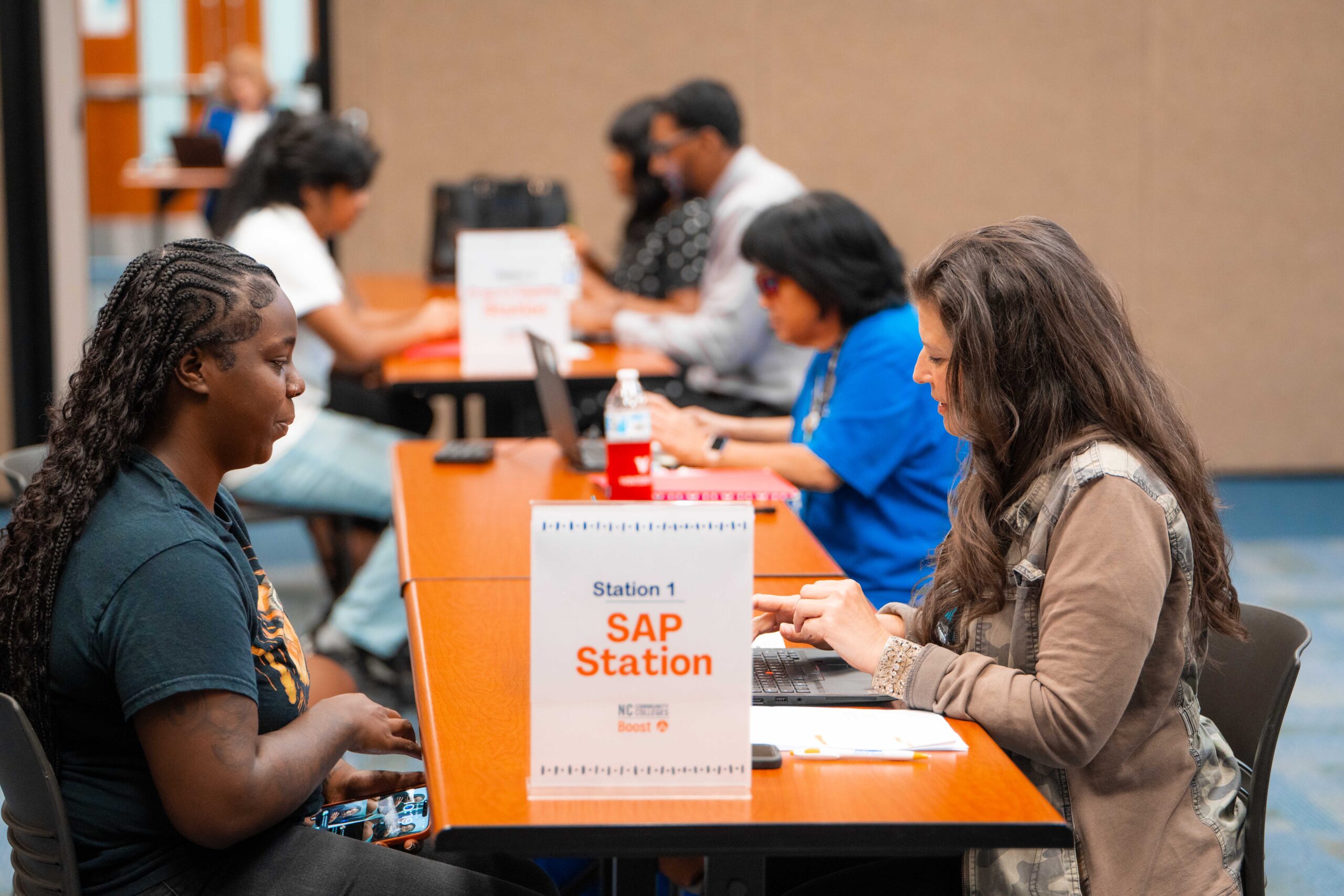 People sit across from each other at a row of help stations in a large room, focused on laptops and paperwork. A sign in the foreground reads “Station 1 SAP Station, NC Boost.” One woman holds her phone while speaking with another person typing on a laptop.