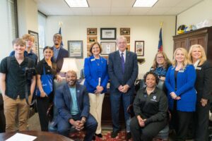 A group of about 12 people, some seated, some standing, all of different ages and races, are in an in door room that looks like an office. Everyone is dressed professionally and is smiling at the camera. 