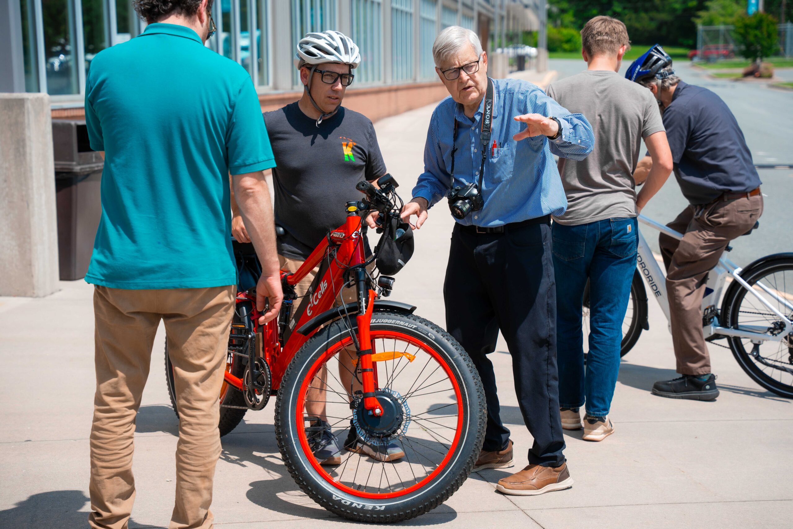 5 men are in the photo, outside in the sun. In the foreground is a man in a teal shirt and khaki pants with his back to the camera. In the center is a man wearing a black polo shirt and a bicycle helmet on a large, red bicycle with a man dressed all in blue with grey hair and a camera around his neck gesturing to something out of the frame. In the background are two more men, with their backs to the camera, with another bicycle. 